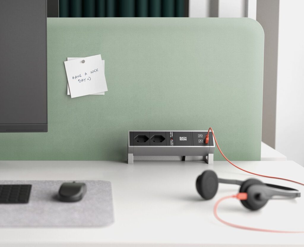 Moderne kantoorwerkplek met een BACHMANN opbouwstekkerdoos inclusief USB-poorten, geplaatst tegen een groen privacyscherm op een wit bureau.