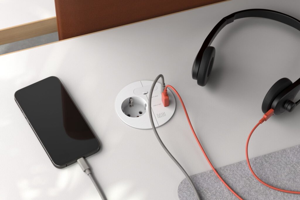 Esthetische kantoorafwerking met een ronde BACHMANN inbouwstekkerdoos in een wit bureaublad, voorzien van een smartphone en hoofdtelefoon die via USB worden opgeladen.